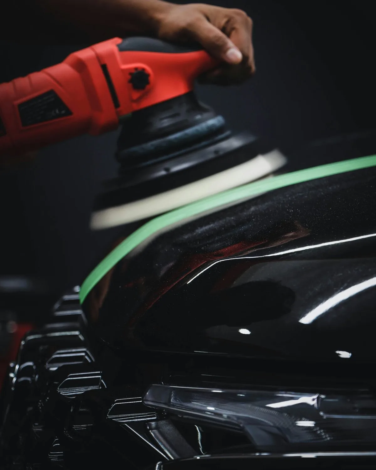 Person using red orbital sander on black car hood with green masking tape visible